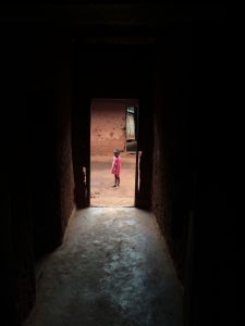 Dark hallway leading to a bright outdoor area, where a small child with pink clothes stands, facing the camera. 